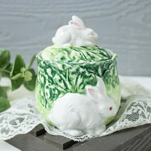 May include: A ceramic jar with a lid, featuring a textured green cabbage leaf design. A white rabbit figurine sits on top of the lid, and a second white rabbit figurine is attached to the front of the jar body. The piece has a glossy finish.