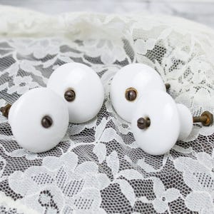 May include: Four round white ceramic knobs with smooth, glossy surfaces. Each knob features a central dark metal screw head and a threaded metal bolt extending from the back, designed for attachment to furniture or cabinetry doors and drawers.