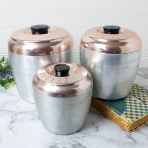 May include: A set of three cylindrical canisters made of brushed aluminum with copper-toned lids and black knobs. Each lid features embossed text reading Flour, Sugar, and Coffee respectively. The containers have a smooth, metallic finish.