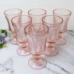 May include: A set of six pink pressed glass goblets featuring a detailed floral and leaf pattern. Each stemmed glass has a circular base and a wide rim, showcasing a translucent light pink hue throughout the entire structure of the glassware.