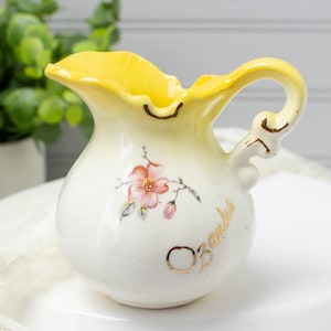 May include: A small, vintage ceramic pitcher with a yellow rim and handle. The pitcher is white with a floral design and the word "Ozarks" in gold lettering. The pitcher has a curved handle and a spout.