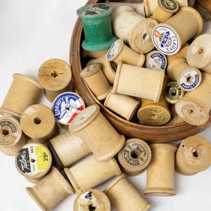 May include: A collection of vintage wooden spools, some with labels from brands like Coats & Clark's and Talon. The spools are various sizes and shades of light brown, with a green spool at the top. Some labels have text like "Liberty" and "Sew-Gude".