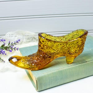 May include: A vintage amber-colored glass shoe, featuring a detailed starburst pattern. The decorative shoe is resting on a light blue book, with a white background and a sprig of purple flowers.