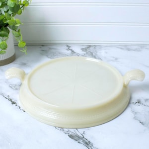 May include: A vintage, off-white, round serving dish with two curved handles. The dish has a decorative rim and a starburst pattern on the interior. The dish is made of a thick, opaque material.