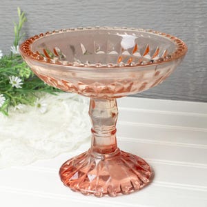 May include: A vintage pink glass pedestal bowl. The bowl has a round, shallow dish with a beaded rim. The stem is a fluted design, and the base is a diamond pattern. The bowl is likely used for serving desserts or displaying decorative items.