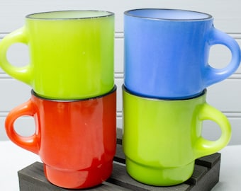 May include: Four colorful ceramic mugs stacked on a dark wooden surface. The mugs are in shades of green, red, and blue, each with a rounded handle. The mugs have a glossy finish and a black trim around the rim.