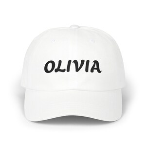 May include: A white baseball cap with the name "OLIVIA" printed in black, cursive lettering across the front. The cap has a curved brim and a button on top.