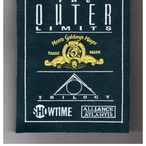 May include: A black fabric patch with white embroidery. The text "THE OUTER LIMITS" is at the top. The Metro-Goldwyn-Mayer logo is in the center with the text "TRADE MARK". Below the logo is a triangle with a circle inside. The text "TRILOGY" is below the triangle. The text "SHOWTIME" is at the bottom left and "ALLIANCE ATLANTIS" is at the bottom right.