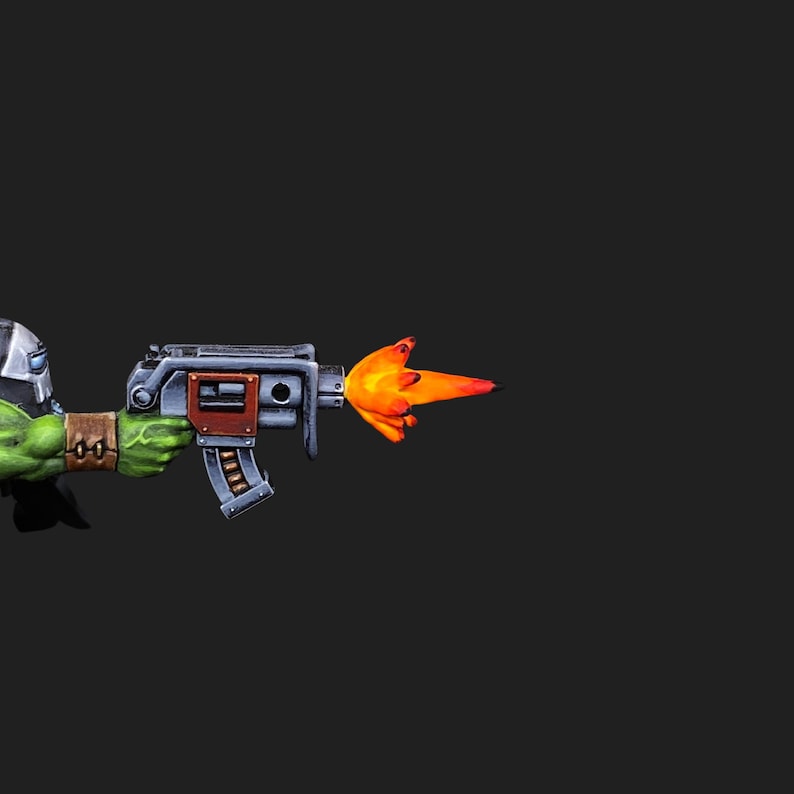 May include: A green figure holding a silver and grey gun with a bright orange blast of fire emitting from the barrel. The figure wears a silver helmet and green wristbands. The background is solid black.