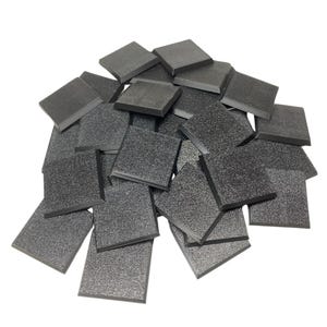 Op de afbeelding: Een stapel kleine, vierkante, donkergrijze tegels met een textuuroppervlak. De tegels zijn opgestapeld en verspreid, waardoor een willekeurige opstelling ontstaat. De randen van de tegels zijn licht afgeschuind.
