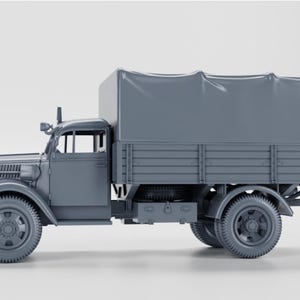 Puede incluir: Maqueta gris de un camión antiguo con una caja de carga cubierta. El camión presenta una cabina, faros y ruedas. La caja de carga está cubierta con un material tipo lona, dándole un aspecto clásico.