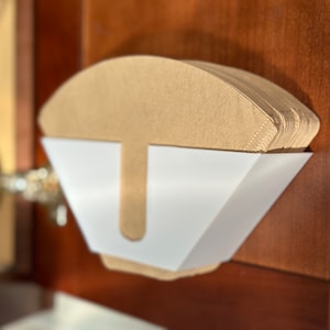 Op de afbeelding: Een witte plastic houder met een stapel bruine papieren koffiefilters erin. De houder is bevestigd aan een houten kastdeur.