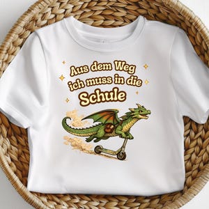 May include: A white t-shirt featuring a graphic of a green dragon riding a kick scooter. The dragon wears a brown satchel. Above the dragon, the text reads "Aus dem Weg ich muss in die Schule" in brown letters, surrounded by small yellow star icons.