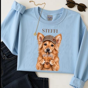 Op de afbeelding: Een lichtblauw sweatshirt met lange mouwen en een afbeelding van een goudkleurige hond met een bruine honkbalpet die een ijskoffie drinkt. De naam STEFFI staat boven de hond. Een donkerblauwe spijkerbroek, een gouden ketting en een zonnebril zijn zichtbaar.