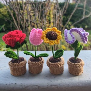 May include: Four crocheted flower pots with different colored flowers. The flowers are a red rose, a pink tulip, a yellow sunflower, and a purple flower. The pots are all brown and have a crocheted texture.