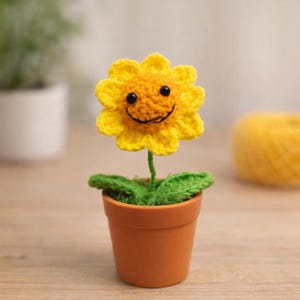 Puede incluir: Un girasol de ganchillo hecho a mano con una cara sonriente, ojos negros y pétalos amarillos. La flor tiene un tallo y hojas verdes, plantada en una pequeña maceta de terracota. Un artículo decorativo alegre.