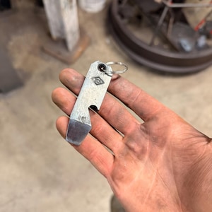 May include: A silver metal bottle opener with a key ring. The opener has a rectangular shape with a beveled edge and a bottle cap opener. The text "BD" is visible on the opener.