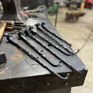 May include: Six hand-forged, black metal hooks with twisted stems and rounded heads are arranged on a metal workbench. The hooks have a curved end. The background shows a workshop with tools and equipment.