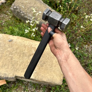 May include: A forged steel hammer with a black handle. The hammer head is a rectangular shape with a textured, metallic finish. The handle is long and straight, with a smooth, dark finish. The hammer is held in a hand.