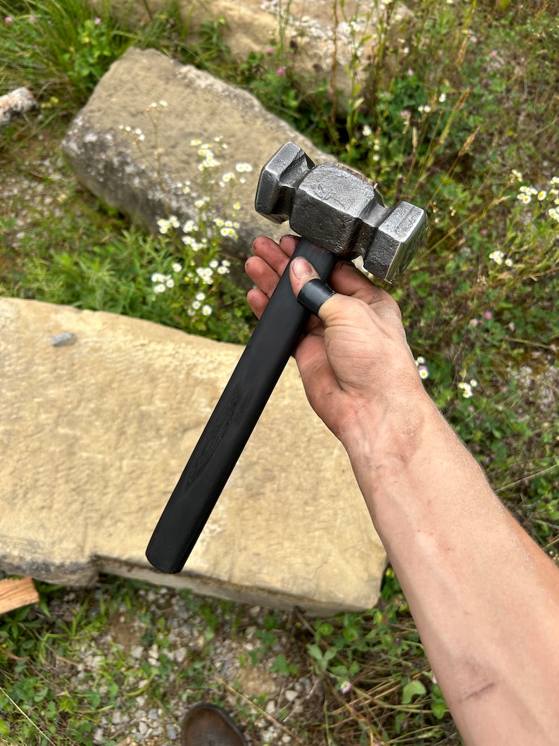 May include: A forged steel hammer with a black handle. The hammer head is a rectangular shape with a textured, metallic finish. The handle is long and straight, with a smooth, dark finish. The hammer is held in a hand.