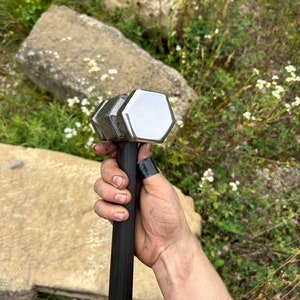 May include: A large, silver and black hammer with a hexagonal head. The hammer's head is polished and reflects light. The handle is black and appears to be made of wood. The hammer is held in a hand.