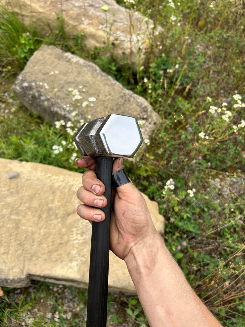 May include: A large, silver and black hammer with a hexagonal head. The hammer's head is polished and reflects light. The handle is black and appears to be made of wood. The hammer is held in a hand.