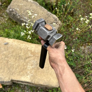 May include: A heavy-duty, gray metal hammer with a black handle and a wooden insert on the head. The hammer is designed for striking and shaping metal. The tool is held in a hand against a natural outdoor background.