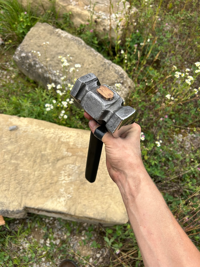 May include: A heavy-duty, gray metal hammer with a black handle and a wooden insert on the head. The hammer is designed for striking and shaping metal. The tool is held in a hand against a natural outdoor background.