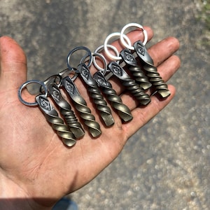 May include: A collection of handcrafted metal keychains. Each keychain features a twisted, bronze-colored design with a silver key ring. The keychains are held in a hand, showcasing their unique, artisan craftsmanship. The keychains have a diamond-shaped design.