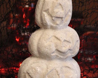May include: A stacked sculpture of three off-white, textured pumpkins with carved jack-o'-lantern faces. The pumpkins are arranged vertically, with the smallest at the top and the largest at the base, featuring a rough, stone-like surface finish.