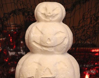 May include: A stack of three off-white, textured pumpkins arranged vertically. Each pumpkin features a carved jack-o'-lantern face with triangular eyes and a wide, smiling mouth. The surface of the pumpkins appears matte and porous in texture.
