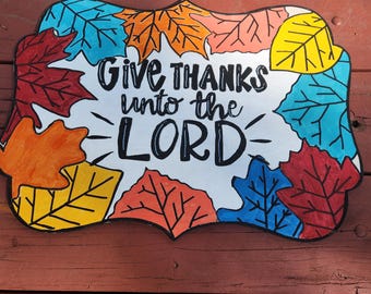 May include: A white sign with the text "Give thanks unto the Lord" surrounded by colorful fall leaves. The leaves are painted in shades of red, orange, yellow, and blue.