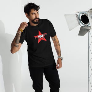 May include: Black t-shirt featuring a red star graphic with the word "RAF" in white. The design includes a stylized image of a weapon. The shirt is made of a soft material.