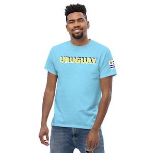 Pode incluir: Uma camiseta azul claro com a palavra "URUGUAY" impressa em letras amarelas com contornos pretos. A camiseta tem um pequeno patch da bandeira uruguaia na manga direita.