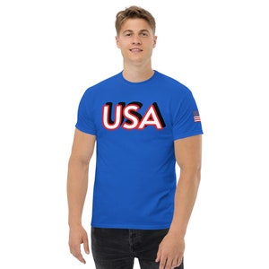 May include: A royal blue t-shirt with a white and red "USA" graphic on the front. The graphic has a 3D effect. A small American flag is on the right sleeve.