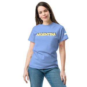 May include: A light blue t-shirt with the word "ARGENTINA" printed in yellow letters with a black outline. The shirt has a small blue and white stripe on the right sleeve.