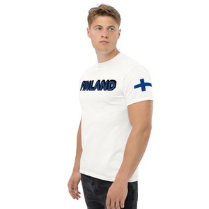May include: White t-shirt with the word "FINLAND" in blue and black lettering across the chest. A blue and white Finnish flag is on the left sleeve. The shirt is made of a soft material.