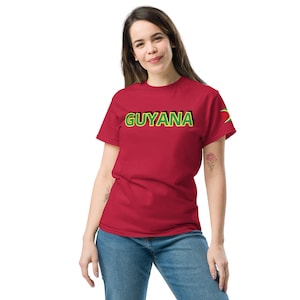 May include: A red t-shirt with the word "GUYANA" in green and black lettering across the chest. The Guyana flag is on the left sleeve. The shirt is made of a soft material and is being worn.