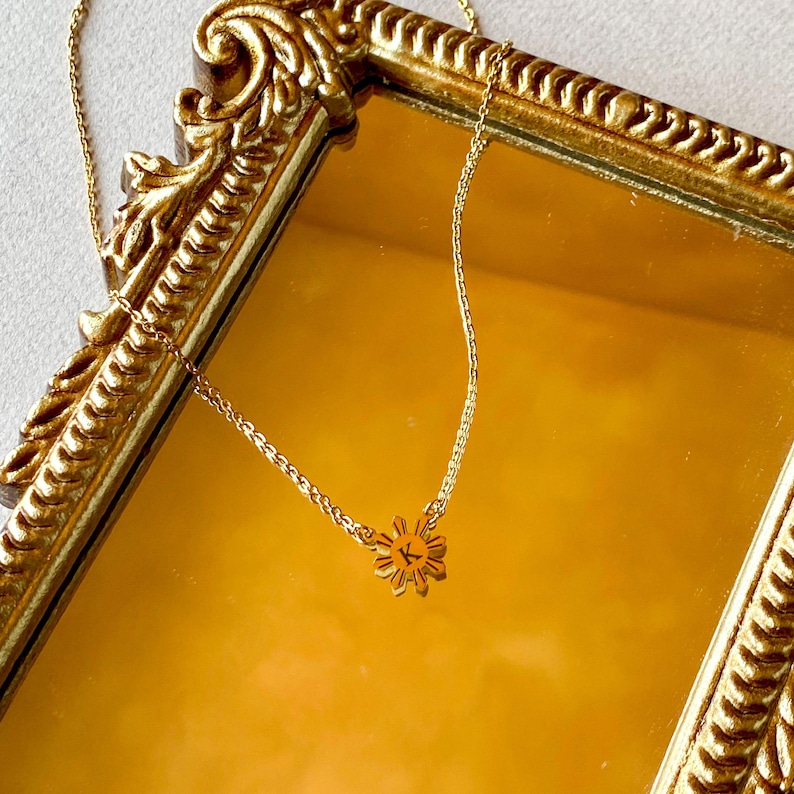 K&ouml;nnte beinhalten: Eine goldene Kettenkette mit einem kleinen goldenen Sonnenanh&auml;nger mit dem Buchstaben "K" in der Mitte. Die Kette wird auf einer goldfarbenen Oberfl&auml;che pr&auml;sentiert.