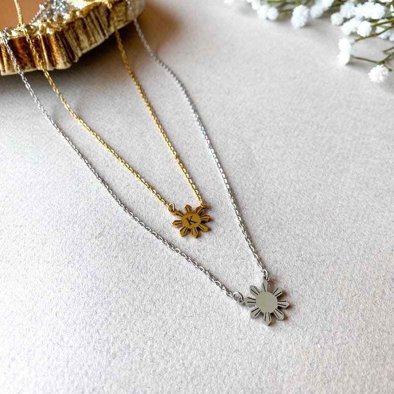 K&ouml;nnte beinhalten: Zwei goldene und silberne Halsketten mit einem Sonnenanh&auml;nger. Die goldene Halskette hat einen kleinen Buchstaben "K" auf dem Sonnenanh&auml;nger. Die silberne Halskette hat einen gr&ouml;&szlig;eren Sonnenanh&auml;nger.