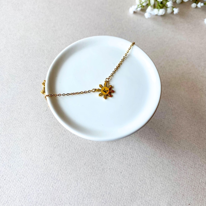 K&ouml;nnte beinhalten: Eine goldene Halskette mit einem Sonnenanh&auml;nger. Der Anh&auml;nger ist eine stilisierte Sonne mit acht Strahlen und einem Buchstaben "K" in der Mitte.
