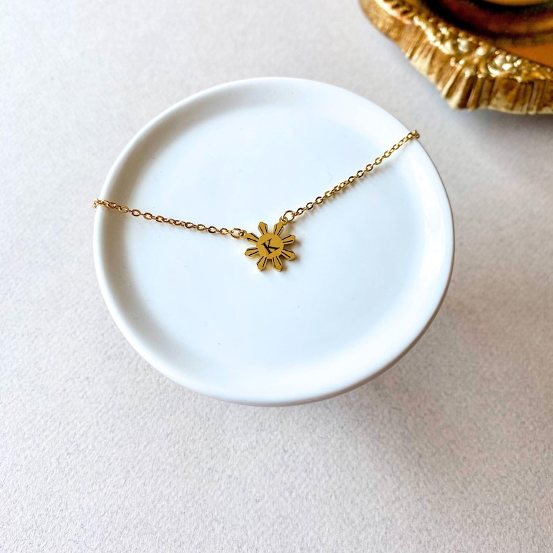 K&ouml;nnte beinhalten: Eine goldene Halskette mit einem Sonnenanh&auml;nger, der den Buchstaben K zeigt. Die Kette liegt auf einem wei&szlig;en Teller.