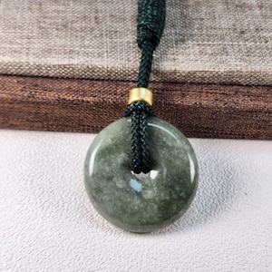 May include: A dark green jade donut pendant with a dark green woven cord. The pendant has a hole in the center and a gold-colored metal accent. The background is a neutral color.