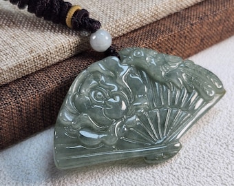 Puede incluir: Un colgante de jade tallado en forma de abanico con un diseño floral. El colgante es verde y tiene un acabado liso y pulido.