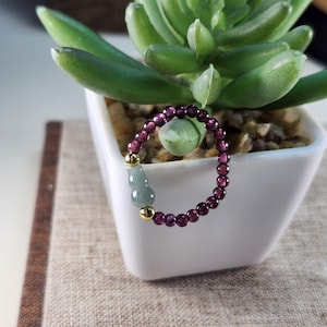 May include: A beaded bracelet featuring deep purple garnet stones and gold accents. The bracelet includes a light green jade pendant. The jewelry is displayed near a succulent plant in a white pot.