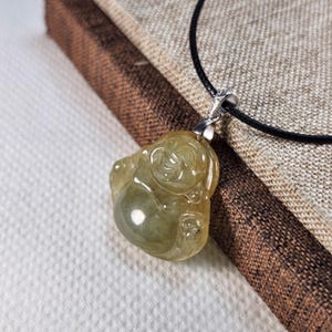 May include: A translucent, light green jade pendant in the shape of a laughing Buddha. The pendant is suspended from a black cord with a silver-colored bail. The Buddha figure has detailed facial features and is set against a neutral background.