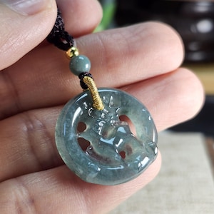 May include: A translucent, circular jade pendant with a carved design, suspended from a black cord. The pendant features a light green hue with darker green accents. A gold-colored accent and a green bead are also visible.