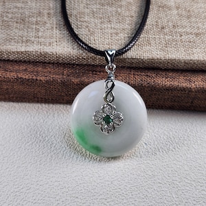 Puede incluir: Colgante redondo de jade blanco y verde con un diseño de trébol plateado, con una piedra preciosa verde en el centro y pequeñas piedras transparentes. El colgante cuelga de un collar de cordón negro.