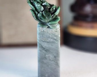 Puede incluir: Una escultura de jade tallada con una flor de loto verde oscuro sobre una base rectangular de piedra gris verdosa. Los pétalos de la flor están detallados y la piedra tiene un aspecto veteado. Esta pieza decorativa es probablemente un pisapapeles o una pequeña escultura.
