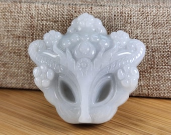 Op de afbeelding: Een witte jade hanger gegraveerd in de vorm van een gezicht met ingewikkelde details. De hanger heeft een delicaat ontwerp met een bloemenmotief en een centraal gezicht met grote ogen.